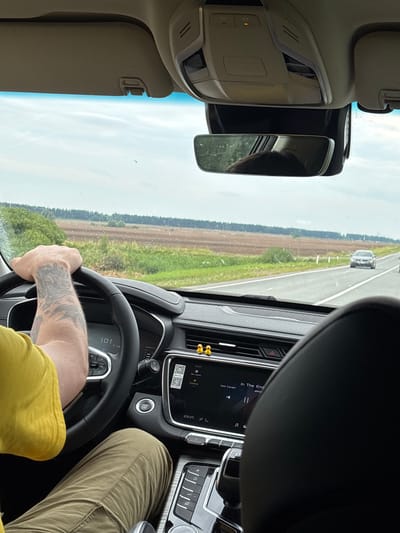 The image shows the interior of a car driving on a highway. The driver's hands are on the steering wheel, and the dashboard is visible, showing the car's speed at 101 km/h. The car's infotainment system is displaying a song titled "In The End" by "soul cover". Outside the car, a rural landscape with fields and trees can be seen. There is a grey car in front of the vehicle. The location is the M5 highway near Shagaban', Russia, as indicated by the highway number and the surrounding landscape. The weather appears to be overcast with light rain. The driver is wearing a yellow shirt and khaki pants.