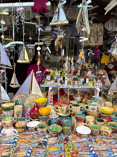 The image depicts a vibrant handicraft market stall, likely located in Abrau-Dyurso, Russia, given the Cyrillic inscriptions on some of the items. The stall is overflowing with a diverse array of handmade decorative items, primarily focusing on maritime and village themes. Suspended from above are numerous small sailboats made from driftwood and fabric, along with whimsical jellyfish-like ornaments adorned with beads and shells.
The central part of the stall features a charming miniature village scene with colorful, tiny houses and lighthouses, crafted from painted wood and ceramic. This is surrounded by a collection of ceramic pottery, including cups, bowls, and plates, many of which are hand-painted with floral motifs, fruits, and nautical elements like fish and lighthouses. Some of the ceramic pieces have small inscriptions in Cyrillic.
In the background, a person with glasses, wearing a pink long-sleeved shirt, stands behind the stall, appearing to be the vendor or a participant in the market. The overall impression is one of a bustling, artisan market filled with unique, handcrafted souvenirs that evoke a sense of coastal charm and folk art. The lighting suggests it is daytime, possibly outdoors or in a well-lit indoor market.