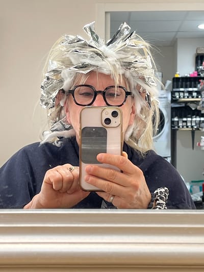 A woman with blonde hair and black-rimmed glasses is taking a selfie in a mirror. Her hair is partially covered with silver foil, suggesting a hair dyeing or highlighting process. She is holding an iPhone with a light-colored case. She is wearing a dark blue top and a patterned scrunchie on her wrist. The background includes a shelf unit with various items and a tiled wall. The environment appears to be a bathroom or salon-like setting. It is not possible to verify the location of the city of Montrond-les-Bains, France based on visual information.
