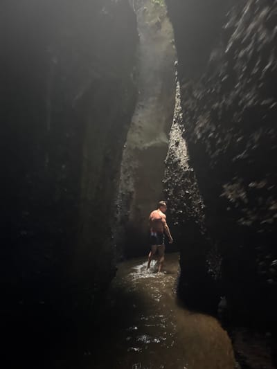 A bare-chested man wearing swim trunks walks through a narrow, dark gorge with water flowing around his ankles. The walls of the gorge are rough and uneven, with some vegetation visible on the upper edges. A shaft of light penetrates from above, illuminating the man and the water. This scene likely depicts Tukad Cepung Waterfall in Bangli, Indonesia, a popular tourist destination known for its unique cave-like waterfall.