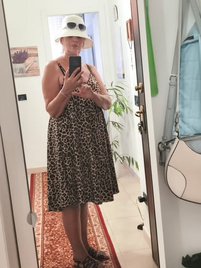 A woman is standing in front of a mirror, taking a selfie. She is wearing a leopard print dress, a white hat with sunglasses perched on it, and dark sandals. Her fingernails are painted bright orange. The background shows a wall with a painting of lavender in a watering can, a door with a key in the lock, a plant, and a light blue quilted jacket hanging on a hook. A patterned rug is visible on the floor. The scene appears to be inside a home, likely in the city of Livorno, Italy.