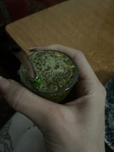 A hand firmly grasps a glass cup filled with a frothy, greenish-brown liquid, resembling a herbal tea or infusion. A metal straw, presumably for sipping, is partially submerged in the liquid. The bubbly surface suggests a freshly poured or brewed beverage. The background shows a blurred table or countertop. The image is close-up, emphasizing the drink and the hand holding it.