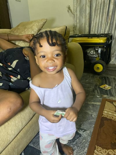A young child with braided hair lies on a beige couch, holding what appears to be money. She is smiling at the camera. The child is wearing a light-colored tank top and patterned pants. An adult arm is visible behind her. In the background, a yellow generator sits near the edge of the couch, and a piece of paper is on a tiled floor near a brown rug. The scene appears to be indoors.