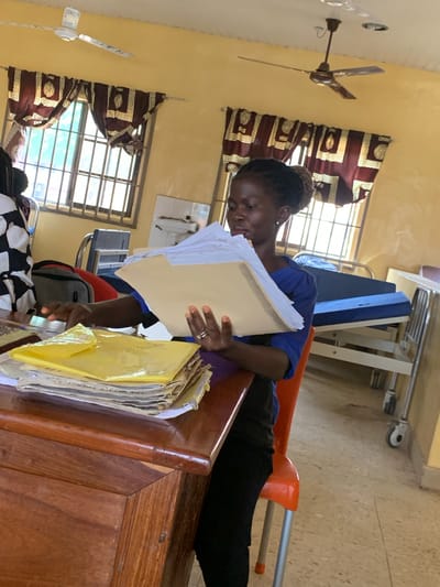 A woman is seated at a wooden desk in a room in Oko Erin, Nigeria. She is reviewing paperwork, holding a large file in her hands. The room features windows with burgundy patterned curtains, a ceiling fan, a bed, and a cart. The woman is wearing a blue shirt and black pants. Additional files are stacked on the desk.