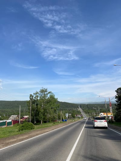 The image depicts a highway leading into a rural area with a clear blue sky and scattered white clouds. On the left side of the road, there are houses with red and green fences, surrounded by lush green grass and trees. Tall power lines run parallel to the road. On the right side of the road, more power lines and streetlights are visible, along with trees. In the distance, a long stretch of highway winds through a valley surrounded by green, tree-covered hills. Several cars are visible on the highway, including a white SUV in the foreground. A billboard advertising "SIBERIAN ZOO" is also present on the right side of the road. The scene appears to be a typical summer day in a semi-rural setting. The location is identified as Bolshaya Rechka, Russia.