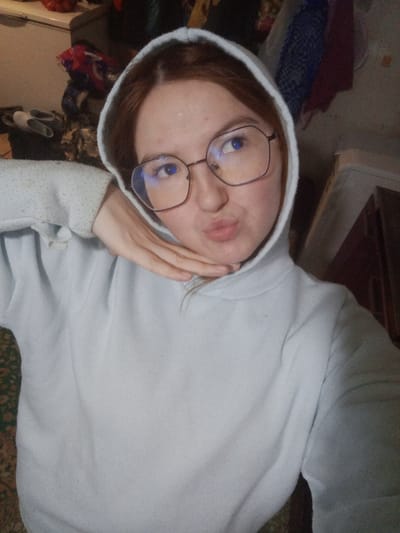 An individual with reddish-brown hair and fair skin is depicted in a close-up, indoor portrait. The subject wears a light grey hooded sweatshirt with the hood up and large, dark-framed, possibly hexagonal glasses, reflecting blue light in the lenses. The subject's lips are pursed into a pout, and the right hand is positioned under the chin. The gaze is directed upwards and to the right. The background shows an indoor environment featuring a patterned rug in the lower left, a pile of various fabrics or clothing items, and a pair of light-colored slippers in the upper left, and dark wooden furniture to the right. The specified location for this scene is Старый Шагирт, Russia.