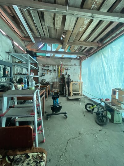 The image depicts the interior of a cluttered workshop, likely a garage or shed, in Tsalyk, Russia. The space is filled with various tools and equipment, suggesting it is actively used for manual labor or repairs.
Dominating the scene is a collection of shelving units laden with items, including bottles of liquid, tools, and containers. A stepladder, partially dismantled, stands prominently on the left. Various hand and power tools are mounted on the wall behind a workbench.
In the center of the workshop, a blue and black industrial pump or compressor is visible. To the right, a small bicycle with yellow handlebars is parked near another workbench. A large, light blue tarp covers a significant portion of the back wall.
The ceiling is constructed from rough wooden planks, and a fluorescent light fixture hangs from the beams. The floor is concrete and appears worn, with some cracks and debris. The overall impression is one of a functional, albeit untidy, workspace. There are no individuals present in the image.