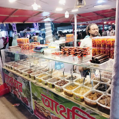 This media file captures a vibrant outdoor food stall, likely at a night market or fair in Charfasson, Bangladesh. The scene is illuminated by overhead lights attached to a temporary red-canopied structure, suggesting it's evening or night.

The central subject is a man with a beard and curly hair, wearing a light-colored t-shirt, standing behind a large glass display counter. He appears to be a vendor or stall operator. To his left, another man and a child in a white cap are partially visible, looking at the display. In the background, numerous people, including women in hijabs, are seen milling about, indicating a busy public gathering. Neon lights and other market stalls are visible in the distance.

The stall prominently features various food items. The glass counter showcases numerous square plastic containers filled with different types of pickles or chutneys ("achar" in Bengali), each with a spoon for serving. These appear to include mango, wood apple, sweet chili, and single-clove garlic variations, as indicated by visible Bengali labels like "আমস" (mangoes), "কদবেল" (wood apple), "মিষ্টি মরিচ" (sweet chili), and "একদানা রসুন" (single clove garlic).

Above the counter, a tiered display of packaged items includes stacks of thin, dark rolls, and numerous transparent plastic jars filled with reddish-orange snacks. These jars have "Natural" and "ন্যাচারাল" (Natural) branding, along with "ASTETO" visible on some labels. More individually wrapped sweet or savory snacks are piled on the counter. A green weighing scale marked "MEA" is also visible.

Banners below the counter display Bengali text. One large banner has "আল্লাহ" (Allah) and "সর্বশক্তিমান" (Almighty), alongside imagery of food. Other text includes "মাছা হাফ বডি ফ্রাই চিংড়ি ঝাল ফ্রাই" (Fish Half Body Fry Prawn Spicy Fry) and "চিকেন ফ্রাই" (Chicken Fry), suggesting other food items might be offered. The overall atmosphere is bustling and lively, typical of a market setting.