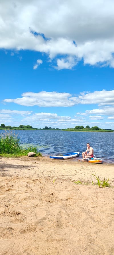 A man is sitting on a paddleboard in the water at the edge of a sandy beach. He is wearing red swim shorts and is facing the camera. His left leg is submerged in the water. Another paddleboard, blue and white, is next to him, partially in the water and partially on the sand. The background shows a wide, calm body of water with a green, tree-lined shore and a bright blue sky with scattered white clouds. There is green grass and a rock on the sandy bank in the foreground. The location is Щиглицы, Russia.