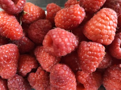 This is a close-up, overhead shot of a pile of ripe raspberries. The berries are a vibrant red and appear fresh, with visible drupelets. The lighting highlights the textured surface of the fruit, and some of the berries show a slight sheen. The arrangement is organic, with berries overlapping and filling the frame. The scene does not depict any identifiable locations in Mieszkowice, Poland, nor does it include any people, actions, or interactions.
