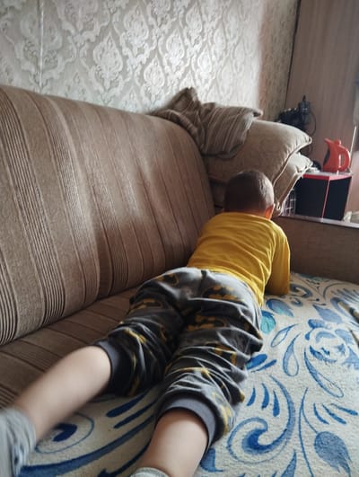 A young child, viewed from behind, is lying prone on a couch. The child is wearing a yellow long-sleeved shirt and patterned gray pants. The couch is upholstered in a brown fabric with vertical stripes. The child's legs are extended towards the viewer. The couch is positioned in front of a wall with textured wallpaper. Behind the couch, there are pillows and a small shelf with an orange kettle and a black electronic device. The scene appears to be indoors, within a residential setting. The image does not provide information to determine the specific location in Kingisepp, Russia.