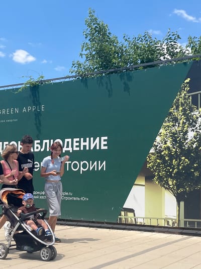 In Yablonovskiy, Russia, on a sunny day, a family walks along a paved path in front of a large green banner. The banner has text printed on it in white, including "GREEN APPLE" and other Russian words. A woman in a pink top and sunglasses pushes a baby stroller with a young child wearing a blue cap. Beside her, a young man in a black Puma t-shirt walks, and a young woman in a light blue t-shirt and grey sweatpants holds a water bottle and a phone. The background shows trees and a building behind the banner.