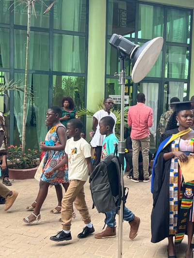 This outdoor scene in Madina, Ghana, depicts a gathering of individuals on a paved area in front of a multi-story building featuring green-tinted glass panels and light green walls. A photography strobe light, specifically a Godox AD600BM with a white beauty dish, is centrally positioned on a silver stand, from which a black backpack hangs. Three children, appearing to be between approximately 6 and 12 years old, walk from left to right across the foreground. The leftmost child, a girl wearing a multi-colored sleeveless dress and glasses, is followed by two boys; one wears a cream t-shirt and tan pants, the other a light blue t-shirt and blue jeans. In the right foreground, an adult woman in a black academic gown with a Kente cloth stole and a black graduation cap holds a light-colored document or certificate. Several adults are visible in the mid-ground: a man in a white short-sleeved top and glasses stands to the left of the light stand, an adult woman in a dark green dress behind him holds a smartphone, and an adult man in a salmon long-sleeved shirt stands with his back to the viewer. A partially visible sign on the building reads "FACE MA" and "ENTR" next to a mask icon, likely indicating entry protocols. Potted plants with pink flowers and a thin palm-like tree are visible on the left side of the frame. The ground is paved with tan interlocking bricks. The scene is illuminated by natural daylight.
