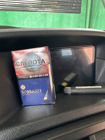 The image shows a close-up view of a car's interior, specifically a storage compartment. In the foreground, a blue and red pack of "Sobranie" cigarettes is visible, displaying the Russian word "СЛЕПОТА" (blindness) prominently. To the right of the cigarette pack, a black electronic device, possibly a stylus or pen, is placed horizontally. A small, rectangular dark screen is also present in the background. The overall setting appears to be a vehicle's dashboard or center console. There is no indication of the city of Nazran, Russia, in the image itself.