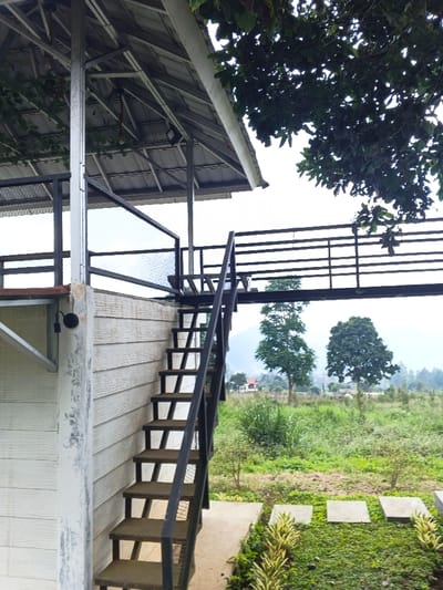 The image depicts a multi-level outdoor structure located in Man Gyat, Myanmar, under an overcast sky. In the foreground, a black metal staircase with approximately ten visible steps and an open riser design ascends towards an elevated platform. The staircase features black metal handrails with horizontal bars. To the left of the staircase, a white building wall with horizontal paneling is visible, featuring a black cylindrical light fixture mounted on its exterior.
Above the first level, a metal-framed structure supports a slanted, corrugated metal roof. An elevated walkway or balcony, enclosed by black metal railings similar to the staircase, extends from the platform. The immediate ground area features a manicured lawn with square concrete stepping stones leading away from the staircase base, flanked by striped green and yellow foliage.
The midground reveals a natural landscape dominated by green grass and scattered bushes. Two prominent deciduous trees with full green canopies stand in the center-right. In the distant background, hazy green mountains or hills are visible beneath the cloudy sky, with hints of other structures in the far-off terrain. No individuals or animals are present in the scene.