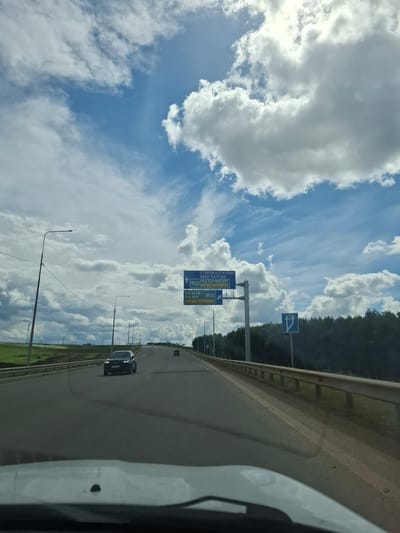 The image depicts a view from inside a vehicle traveling on a multi-lane highway under a cloudy sky. The highway is paved and has visible lane markings. In the distance, other vehicles are seen on the road. On the right side of the highway, there is a dense line of trees, and on the left, a grassy embankment. Overhead, large, white cumulus clouds are scattered across a blue sky, with some wispy cirrus clouds also present.
Road signs are visible on the right side of the highway. One sign is blue with white text, indicating directions to "СТАРОБАЛТАЧЕВО", "ИСК БАЛТАС", "ЕКАТЕРИНБУРГ", and "M12 ПЛАТНАЯ ДОРОГА". Another smaller blue sign with white arrows indicates a lane split. A blue sign with a white arrow pointing upwards and to the right is also visible.
The location is identified as the city of Уразгильды, Russia, specifically on a highway.