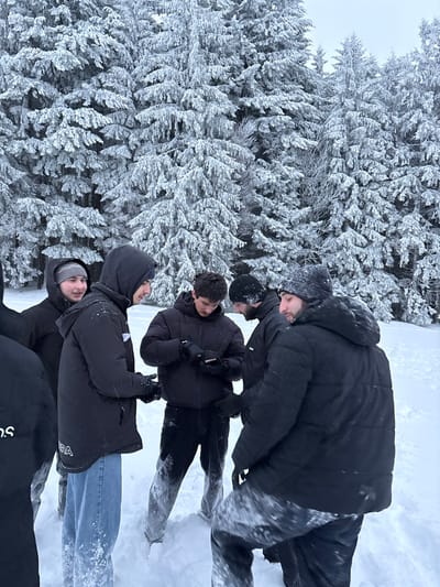 Six males are gathered in a snow-covered clearing within a dense fir forest in Le Hohwald, France. The forest and ground are heavily covered in white snow and frost under an overcast sky. All individuals wear winter attire, including dark puffer jackets, hoodies, beanies, and gloves. Snow is visible on their clothing, particularly on the lower portions of their pants and jackets. One male in the center is actively observing and interacting with a smartphone held in his gloved hands. Another male to his immediate left is looking towards the smartphone user. A third male to the right of the smartphone user is also partially obscured but appears to be looking towards the device. A fourth male on the far left, behind the second male, is smiling and looking towards the center of the group. Two other males are partially visible on the far left and far right. The group is positioned in a loose cluster, indicating a collective interaction or observation.