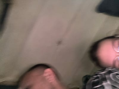 This is a blurry, low-resolution, close-up photograph taken from a low angle, appearing to be an interior shot. The primary subjects are two individuals, their faces obscured by motion blur and poor lighting. One person's head is visible in the foreground on the left, showing a pinkish tone and what might be a hand or finger. Another person's head is visible on the right, with a pinkish complexion and what could be glasses or a light reflection on their cheek. They are both wearing patterned clothing. The background is a muted, textured surface, possibly a ceiling or wall, with some darker shadows. There are no discernible objects, text, or clear cues about the time of day, weather, or specific location within Kyrgyzstan. The overall impression is that of a candid, quickly captured moment, perhaps a selfie or a snapshot of people in close proximity.