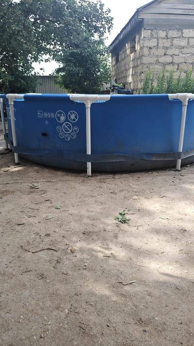 An outdoor scene in Индербор кенттік әкімдігі, Kazakhstan, features a blue above-ground swimming pool situated on dry, sandy ground. The pool is supported by white vertical poles and has black strapping around its base. Several circular warning symbols are visible on the blue exterior of the pool. In the background, a building constructed with light-colored bricks is partially visible, along with lush green trees and foliage. The ground is uneven and scattered with small stones and fallen leaves. The lighting suggests it is daytime, with shadows indicating the presence of overhead trees.