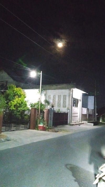 This media file depicts a nighttime street scene in Julicotmeurak, Indonesia. The main subjects are a residential building and its surroundings, including a street, a fence, and lush vegetation.

The building appears to be a modest house with a light-colored facade. It features decorative, rectangular window openings and a covered porch area. A dark-colored gate and a black metal fence suggest a boundary around the property, with a gate leading to a pathway or driveway. In front of the fence, there is a vibrant green tree, illuminated by a street lamp.

The street in the foreground is paved and appears damp, possibly from recent rain or dew. A street lamp stands tall on the right side of the image, casting a bright light over the scene. The sky above is dark, characteristic of nighttime, with a prominent full moon partially obscured by clouds. The presence of overhead wires suggests an urban or suburban environment.

The overall mood of the image is quiet and serene, typical of a peaceful night. The moonlight and streetlamp create a soft illumination, highlighting the textures of the buildings and vegetation. There is no visible text in the media file. The scene captures a moment of stillness in a residential area under the glow of the moon.