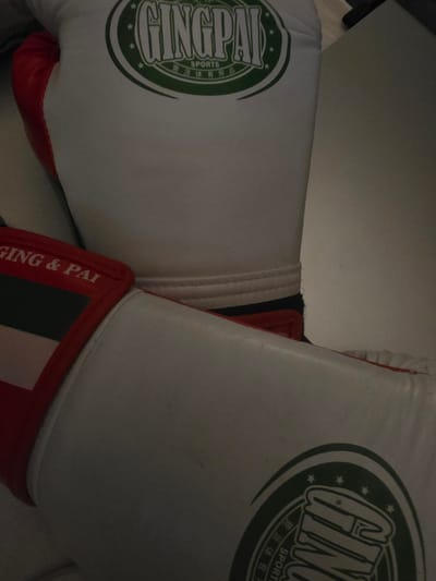 The image shows two white boxing gloves with red accents and green logos. One glove is positioned on top of the other, with its logo prominently displayed. The logo is a circular emblem with the word "GINGPAI" in large, stylized letters, surrounded by smaller text and stars. The other glove, partially visible, has a red band with "GING & PAI" written on it in white letters. The gloves appear to be made of leather. The background is a light-colored, out-of-focus surface, possibly a table or a wall. The lighting is dim, casting shadows on the objects. The scene does not provide any information that would allow for the localization of the image to Dupnitsa, Bulgaria, or any other specific city. There are no people, actions, or interactions visible in the image.