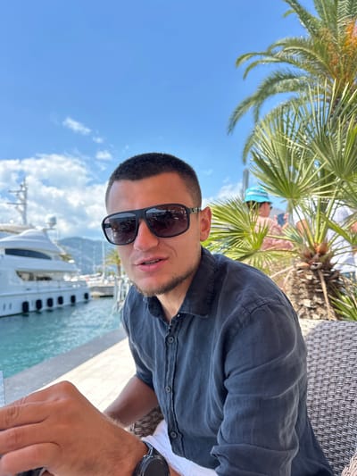 The image shows a man wearing sunglasses and a dark shirt, seated at a table outdoors. Behind him, a large white yacht is docked in a body of water. Palm trees and mountains are visible in the background under a bright blue sky with scattered clouds. The location is identified as Donja Lastva, Montenegro. The man is looking towards the camera with his mouth slightly open, and his left hand is resting on the table. There are other people in the background, partially obscured by palm fronds. The overall scene suggests a leisure setting, possibly a cafe or restaurant by the water.