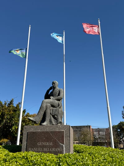 A bronze statue labeled "GENERAL MANUEL BELGRANO" stands on a stone pedestal against a backdrop of green foliage. Behind the statue, three flagpoles display flags against a clear blue sky. The top flag is red with faded white text or symbols. The middle flag is light blue and white. The bottom flag is green with yellow and red elements. In the background, a building with brick accents is visible. The photograph is taken outdoors in the city of Tigre, Argentina.