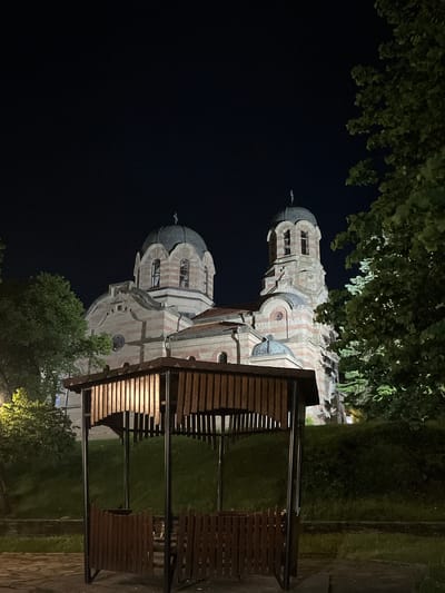 The image depicts the Church of Saints Peter and Paul in Veliki Preslav, Bulgaria, at night. The church, a prominent structure in the midground, features a distinctive striped facade composed of alternating bands of light-colored stone and reddish-brown brick. Two main domes are visible: a larger central dome to the left and a smaller dome to its right. A taller bell tower is situated on the right side of the church, also exhibiting the striped pattern and featuring arched openings. All visible domes and the bell tower are topped with Orthodox crosses.
In the foreground, a hexagonal wooden gazebo occupies the lower-middle portion of the frame. It has a dark, possibly metal, structural frame and walls constructed from vertical wooden slats. An internal light source illuminates the interior of the gazebo and the inner surfaces of its wooden slats, casting a warm glow. No individuals are present within or around the gazebo.
Trees with dark green foliage flank the church on both sides, with a dense cluster of trees and bushes partially obscuring the right side of the church and extending into the background. Another tree is visible on the far left. The ground immediately surrounding the gazebo appears to be paved with stones, transitioning to a grassy slope behind it. The sky above is dark, indicating nighttime, with no celestial bodies visible. Artificial lighting illuminates the church's facade and the gazebo, contrasting with the dark surrounding environment.