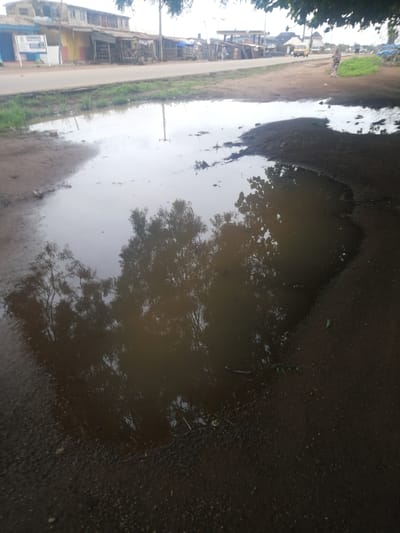 The image shows a large, muddy puddle on a dirt road in Omupo, Nigeria. The puddle reflects the surrounding trees and sky. In the background, there are buildings, a road with some traffic, and a few people walking. The scene appears to be an outdoor environment, possibly a street or a village road, during daytime. The overall impression is one of a rural or semi-urban setting with signs of everyday life.