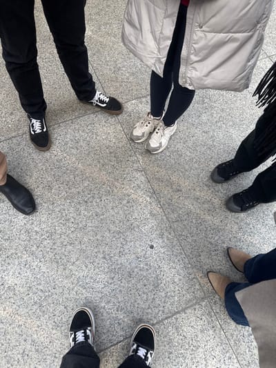 The image depicts the lower extremities of six individuals standing in a rough circle on a light gray speckled granite tile pavement, indicative of an outdoor public space in Visp, Switzerland.

From a high-angle perspective, the visible subjects include:
1.  **Top-left:** A person wearing black trousers and black Vans Old Skool sneakers with white laces and white side stripes, featuring brown gum soles.
2.  **Top-right:** A person wearing black leggings and light beige/white athletic sneakers with textured soles. A beige puffer coat partially covers the legs.
3.  **Mid-right:** A person wearing dark pants and dark (possibly black canvas) low-top sneakers with dark laces and a dark rubber toe cap/sole edge. A black fringed garment is visible beside the legs.
4.  **Bottom-right:** A person wearing blue jeans and light brown/tan pointed-toe heeled boots. A light beige coat is partially visible at the bottom edge.
5.  **Bottom-center:** A person wearing black trousers and black and white checkered Vans Old Skool sneakers with white laces and white side stripes, featuring brown gum soles.
6.  **Mid-left:** A single foot, clad in a black pointed-toe leather flat shoe, with a light brown/tan pant leg visible.

All individuals appear stationary, facing inwards, suggesting a paused social interaction or gathering. The lighting is even, casting minimal shadows.