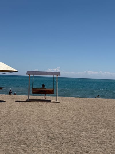 This is a vertical, eye-level, medium shot of a beach on a sunny day. The sky is a clear, light blue, with a few wispy white clouds near the horizon. The sea is a deep turquoise color and appears calm, with small ripples on the surface. A sandy beach occupies the foreground and midground.

In the midground, a wooden swing set with a roof is positioned on the sand. A single person, viewed from the back, is seated on the swing, wearing headphones and facing the water. Their legs are extended forward. To the left of the swing set, two people are visible near the water's edge; one appears to be wearing a dark top and head covering, and the other is a child wearing a blue hat. A portion of a light-colored beach umbrella is visible on the far left.

In the background, further out in the water, a person can be seen swimming. The overall atmosphere is one of a relaxed summer day at the beach. The location is identified as Koshkol', Kyrgyzstan, implying a setting likely on the shores of Issyk-Kul Lake.