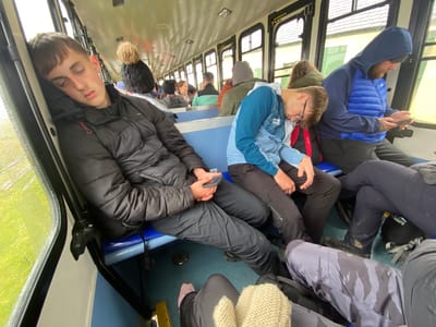 The image depicts the interior of a crowded train carriage, likely the Snowdon Mountain Railway, given the location is Llanberis, United Kingdom. The carriage is filled with passengers sitting on blue seats. A young man in a black puffer jacket is asleep, leaning against the window. He is holding a smartphone in his hand. Next to him, another young man with glasses is also asleep, leaning forward with his head in his hands. To the right of him, a man in a blue hooded jacket and puffer vest is using a smartphone. Other passengers are visible further down the carriage. The window shows a view of green foliage, likely indicating the train is traveling through a natural environment. The light inside the carriage is subdued.