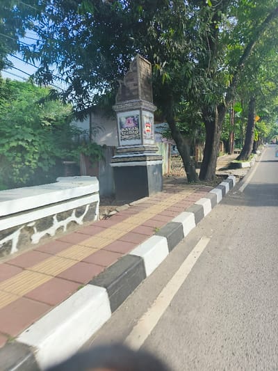 The image shows a sidewalk along a paved road in Purwakarta, Indonesia. The sidewalk is made of red and beige rectangular bricks with white and black borders. A concrete barrier runs along the edge of the road. A large concrete pillar with a sign reading "BATAS KELURAHAN WANAKARTA" is visible in the image. The pillar is partially obscured by a large green tree. The road is empty and appears to be relatively narrow. The sky is blue with a few white clouds. There is a small bird on the sidewalk in front of the pillar.