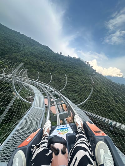 An extreme close-up POV shot shows a person riding a roller coaster down a steep, forested mountainside in Qingyuan City, China. The roller coaster track, made of metal with safety netting on either side, curves sharply to the left as it descends. The person's legs are visible in the foreground, clad in black and white zebra-print pants and white sandals. Their feet are resting on the roller coaster cart, which is gray and orange. The background is dominated by lush green trees covering the mountainside, with a cloudy blue sky above. A black camera is mounted on a post to the right, suggesting this is a popular tourist attraction.