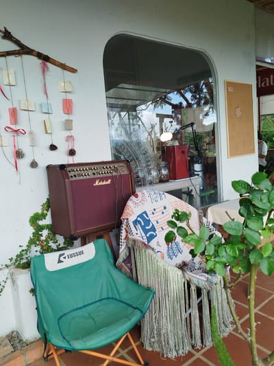 A maroon Marshall amplifier sits on a stand next to a green folding chair. The amplifier is in front of a white wall with vines growing on it. Behind the amplifier is a window that looks into a room with a red coffee maker and a lamp. A blanket with fringe is draped over a chair next to the amplifier. A branch with small paper decorations hangs on the wall above the amplifier and chair. The scene is outdoors, on a tiled patio, with a plant in the foreground. The location is in Trường An, Vietnam.