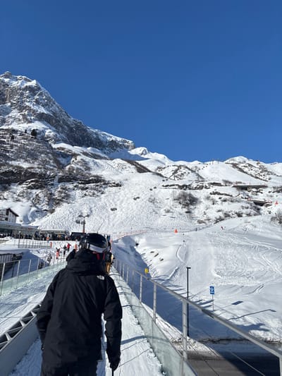 The image presents a snow-covered alpine ski resort in Zürs, Austria, under a clear, bright blue sky. In the immediate foreground, a person is seen from the back, wearing a black ski helmet with white and red goggle straps, a black ski jacket displaying a white "The North Face" logo on the left shoulder, and black gloves. This individual stands on a horizontal moving walkway or conveyor lift, which features translucent glass and metal railings.
To the left of the walkway, a ski lift base station is visible, with a substantial queue of skiers dressed in various colored ski apparel. Adjacent to the lift station is a multi-story building, partially covered in snow. Beyond the moving walkway, expansive ski slopes dominate the midground, crisscrossed with numerous ski tracks. Multiple ski lifts, consistent with chairlifts, ascend the snow-covered mountainsides. Orange conical markers are intermittently placed on the slopes.
In the background, imposing, snow-capped mountains rise steeply, exhibiting both smooth snowfields and rugged rocky outcrops. Sparse trees or brush can be observed on the lower mountain slopes. The overall scene is brightly lit by direct sunlight, indicating active daytime operations at the ski resort.