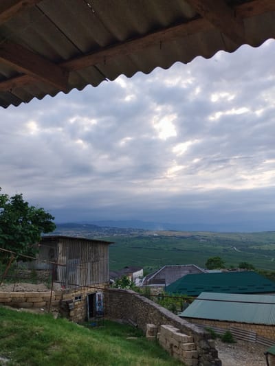 The image depicts a rural landscape with buildings and a cloudy sky. In the foreground, a corrugated metal roof juts into the frame from the top. Below the roof, a rustic wooden structure with a corrugated metal siding stands on a hillside, with a stone wall in front of it and overgrown green grass. To the right of this structure, a path leads past other buildings with green roofs. In the mid-ground, a valley unfolds with scattered houses and agricultural fields, leading to distant mountains under a dramatic sky filled with broken clouds, some illuminated by sunlight. The scene appears to be located in the village of Darvag, Russia, given its mountainous terrain and architectural style. There are no people visible in the image.