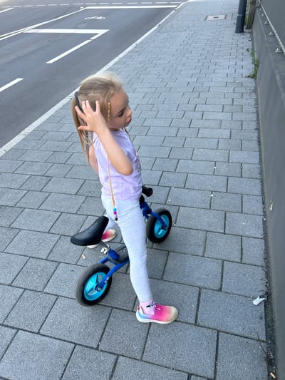 A young Caucasian girl with blonde hair tied in a ponytail with a decorative string of beads is standing next to a blue balance bike on a gray brick sidewalk. She is wearing a light purple t-shirt with a graphic print, light gray leggings, and multi-colored athletic shoes. Her left hand is raised to her hair. The balance bike has a black seat and blue wheels with black tires. The sidewalk is adjacent to a street with white lane markings and a bicycle symbol, and a concrete wall with a metal railing runs along the right side of the sidewalk. The environment appears to be outdoors in a German city, possibly Waldbröl, given the street markings and architecture.