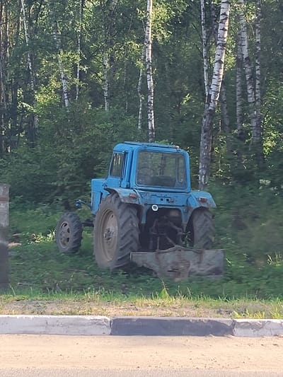 The image shows a blue tractor parked in a grassy area with trees in the background. The tractor is facing away from the viewer, and its cab is visible, as are its large rear wheels and smaller front wheels. A piece of farm equipment, possibly a plow or bucket, is attached to the rear of the tractor. The tractor appears to be old and weathered.
The scene is set outdoors, with a dense forest of birch trees behind the tractor. The ground is covered in green grass and some patches of dry vegetation. In the foreground, there is a paved area with a curb separating it from the grassy area. The lighting suggests it is daytime, possibly late afternoon or early evening, with sunlight filtering through the trees. The overall impression is one of a rural or semi-rural setting in Medik, Russia. There is no text visible in the image. The mood is peaceful and still, with the tractor appearing to be parked for a break.