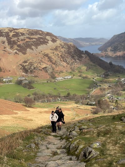 An outdoor scene depicts three individuals hiking uphill on a rocky, stepped path, overlooking the village of Glenridding and Ullswater lake in the United Kingdom. The foreground features a steep, ascending path composed of irregular stone slabs and boulders, bordered by dry brown grasses and scattered green patches. Three people are positioned in a single file on the path, moving upwards. The lead individual, a female, wears a light-colored hooded top, dark trousers, and sunglasses. Behind her are two males, both wearing dark hooded tops and sunglasses; one is in dark shorts and carries a backpack.

Below the path, the midground opens into a valley containing green pastoral fields segmented by dry stone walls. Clusters of traditional stone houses with grey roofs are visible throughout the valley, indicating the village of Glenridding. A narrow road winds through the village. Further into the background, the dark blue waters of Ullswater stretch between rugged, heather-covered fells and hills. A small vessel is discernible on the lake's surface. The sky overhead is partially cloudy with patches of blue, under bright daylight conditions.