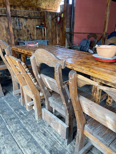 The image shows a cluttered outdoor space, possibly a patio or storage area, in Картмазово, Russia. There is a large, heavily varnished wooden table with items on it, including bowls. A pile of wooden chair parts or disassembled chairs is stacked next to the table. In the background, there's a brick wall, a wooden wall, a fire extinguisher, tools hanging on the wall and some additional kitchenware items stored in the shelves. The flooring is made of weathered wooden planks.