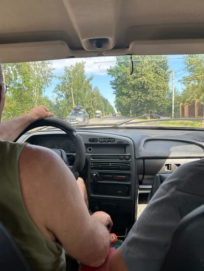 The image depicts the interior of a car driving down a road in Лотошино, Russia. The viewpoint is from the passenger seat, looking towards the driver and the road ahead through the windshield. The driver, a male with his arm on the steering wheel, is wearing a green tank top. A second passenger is visible in the right side of the frame, wearing a grey shirt. The car's dashboard is visible, with instruments and a radio. Outside the car, trees line the road, and a few other vehicles are seen in the distance. The weather appears to be sunny.