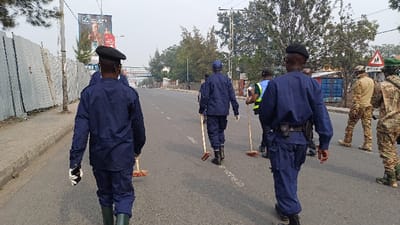 A group of approximately seven individuals is depicted walking forward on a paved two-lane road in Rubavu, Rwanda, during daytime under an overcast sky. The scene appears to be a street cleaning operation.

Five individuals are wearing dark blue uniforms, consisting of long-sleeved jackets and trousers. Three of these individuals wear black berets. One individual in a blue uniform on the far left wears green rubber boots and white gloves, holding a black mobile phone in his left hand. The central figures in blue uniforms wear black boots and hold brooms with wooden handles and red/brown bristles, positioned to sweep the road. One uniformed individual has a holster on their right hip. One person, positioned towards the center-right, wears a yellow/green reflective vest over a dark shirt and also holds a broom. To the far right, two individuals wear military-style camouflage uniforms, with one wearing a tan-colored shirt and camouflage trousers, and the other fully in camouflage, both carrying what appear to be backpacks.

The road features a faint white dashed center line and partially visible white text on the asphalt, possibly reading "CLEAN." On the left side of the road, a concrete sidewalk runs alongside a corrugated metal fence or hoarding. A utility pole is situated along this fence line. In the background on the left, a large billboard with advertisements, including the MTN logo and a male figure, is visible. On the right side of the road, blue shipping containers are present behind the individuals. Further down the road on the right, a triangular traffic sign with a red border and an image of two children indicates a school zone, with the word "ECOLE" partially visible below it. An additional circular green sign is adjacent to the school sign. Sparse trees line parts of the street and appear in the background.