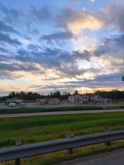 The image shows a cloudy sky at sunset over a commercial area in Rigaud, Canada. The sky is filled with dramatic clouds, with areas of orange and yellow light breaking through, suggesting the sun is low on the horizon. In the foreground, a metal guardrail runs horizontally across the bottom of the frame. Beyond the guardrail is a grassy embankment and a paved road.

Further back, across the road, is a collection of buildings. On the left, a Tim Hortons restaurant is visible, with a white van parked in front of it. To the right of Tim Hortons, there is another commercial building with outdoor seating. Further to the right is a larger, multi-story building that appears to be a hotel or office building, with several windows and a light-colored facade. Trees and vegetation form a backdrop behind the buildings. The overall scene depicts a twilight view of a roadside commercial strip.