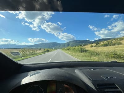 This media file is a first-person perspective shot from inside a car, looking out through the windshield at a scenic rural landscape. The scene depicts a paved road stretching into the distance, flanked by green, grassy fields and rolling hills covered in trees. The sky is a vibrant blue, dotted with fluffy white clouds, suggesting a bright and pleasant day. The time of day appears to be daytime, likely late morning or early afternoon, given the quality of light and shadows.

The foreground is dominated by the car's dashboard and parts of its interior, including a textured surface and what appears to be a car's instrument cluster with some visible text, though it is largely obscured and difficult to decipher. The dashboard is dark-colored and textured, with some reflections from the outside.

The activity taking place is a car journey. The road ahead indicates forward movement through the landscape. The overall mood conveyed is one of peaceful travel and enjoyment of nature's beauty. There are no people clearly visible in the shot, though the perspective implies a driver and possibly passengers are present.

Notable details include the quality of the sky and the lush greenery, which suggest a location with a temperate climate during a favorable season. The presence of a road and the car interior firmly place the scene in a context of modern travel. The location is identified as Orizari, Bulgaria. No specific emotions are overtly expressed, but the imagery evokes a sense of calm and openness. The text on the instrument cluster is not legible enough to be transcribed.