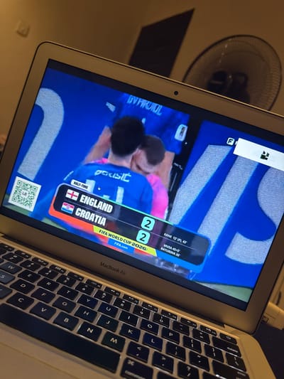 The image shows a laptop screen displaying a soccer match score. On the screen, it shows "HALF-TIME" with England and Croatia tied at 2-2. The game is identified as "FIFA WORLD CUP 2026". Below the score, there are names of players and the times they scored: "KANE 12' (P), 42'", "MUSA 45+5'", and "BATURINA 36'". The laptop is a MacBook Air, and the keyboard is visible in the foreground. A QR code is displayed on the left side of the screen. The background shows a fan and a wall, suggesting an indoor setting. The specific location in Kufena, Nigeria is not discernible from the image.