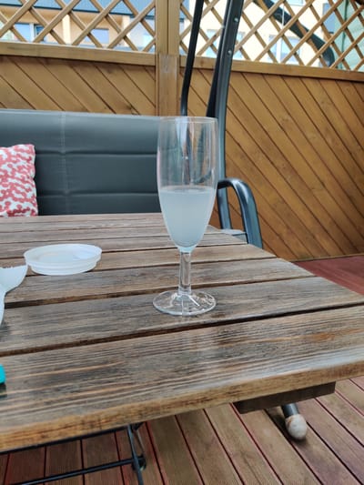 A close-up, eye-level shot shows a wine glass filled with a pale, translucent liquid on a weathered wooden table. The table is made of planks with visible grain and some signs of wear. To the left of the glass is a white plastic lid, possibly from a container, and a crumpled white paper napkin or cloth. In the background, a dark gray cushioned bench is partially visible, with a pillow featuring a red and white pattern. Behind the bench, a wooden lattice fence separates the patio area from an apartment building with windows. The scene is set on a wooden deck, likely a balcony or patio, in Haapsalu, Estonia. The lighting suggests it is daytime, possibly late afternoon.