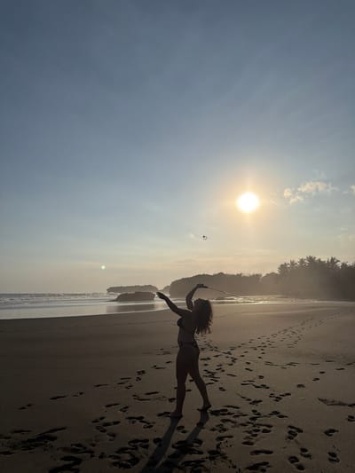 The image depicts a beach scene at either sunrise or sunset in Selemadeg, Indonesia. A single individual, appearing as a dark silhouette, stands on the sandy shore. The person, identified as female with curly hair and wearing a two-piece swimsuit (bikini), is positioned with a badminton racket raised overhead. A white shuttlecock is visible in the air, slightly above and to the left of the racket, indicating an action related to playing badminton, possibly a serve or hit.

The sun is low on the horizon, prominent in the upper right quadrant of the image, casting a strong backlight and creating lens flare. Its intense glow highlights the edges of clouds and a distant line of palm trees on the right side of the frame, which are also silhouetted. The beach consists of dark sand, marked with numerous footprints leading away from the individual. Ocean waves are visible breaking near the shore on the left. In the background, several large rock formations or cliffs emerge from the water and coastline. The sky exhibits a gradient of light blue, transitioning to warmer tones near the sun, with a few scattered clouds.