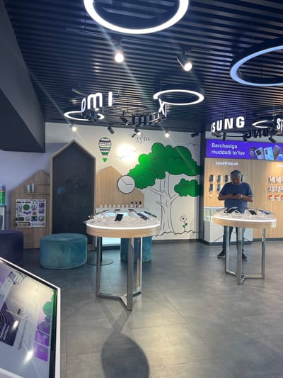The image shows the interior of a modern electronics store, likely a Samsung or Ucell branded retailer, located in the city of Kashgar mahalla, Uzbekistan. The store features a dark, slatted ceiling with prominent circular and linear LED lighting fixtures. White illuminated signage with stylized lettering, possibly brand names, is visible on the ceiling.
The walls are white and decorated with a black and white mural depicting a stylized tree with green foliage, a hot air balloon, and other whimsical elements. A large wooden-framed blackboard or display area is positioned against one wall. To the left, there's a display featuring various electronic accessories and children's toys, with Ucell branding evident.
In the center of the store, there are display tables showcasing mobile phones. A man, wearing a blue t-shirt and jeans, is standing at one of these tables, looking down at a smartphone he holds in his hands. He appears to be browsing or interacting with the device. Nearby, there are round, teal-colored ottomans.
A large digital display screen is visible in the foreground on the left, displaying colorful graphics and text. In the background, on the right, a section of the store is dedicated to "SMARTFONLAR" (smartphones) and "AKSESSUAR" (accessories), with various phone models and other electronic items displayed on shelves. The flooring is a dark gray tiled surface. The overall impression is a bright, contemporary retail environment.