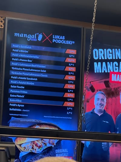 The image shows a menu board at a restaurant called "mangal" located in Köln, Germany at the coordinates 50.93, 6.939. The menu board is predominantly black, with a red and white logo displaying the restaurant name and a red cross, with the word "DONER" written underneath.  The menu lists different food items available, including "Poldi's Sandwich," "Poldi's Durüm," "Poldi's Weltmeisterteller," "Poldi's Pomme-Box," "Poldi's Lahmacun Beef/Chicken," "Türkische Pizza/Lahmacun Salat," "Türkische Pizza/Lahmacun Classic," "Poldi's Falafel Sandwich," "Poldi's Falafel Dürüm," "Salat-Tasche," "Pommes Frites," "Extra Fleisch," "Extra Käse," "Poldi's Ayran," "Softdrinks + Kiosk-0,33l," and "Softgetränke + Kiosk-0,5l," with corresponding prices listed next to each item. Additionally, a portion of the menu shows the price for "Allegetränke (außer Softdrinks) im Restaurant sind frei erhältlich." Below the menu, there is a picture of a kebab sandwich. Above the menu board, a man is seen smiling into the camera, wearing a dark blue shirt. The upper right portion of the image shows a chain-link fence, with the restaurant name and the word "ORIGINAL" appearing in red text on the side. The background consists of a red wall with a white text label saying "mangal" in red lettering, appearing partially in the image. The image is captured in a bright, well-lit indoor environment, with the menu board as the focal point.